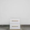 Front view of a modern white bedside table with two drawers, gold handles, and a gold steel base, set on a grey tiled floor against a white wall.