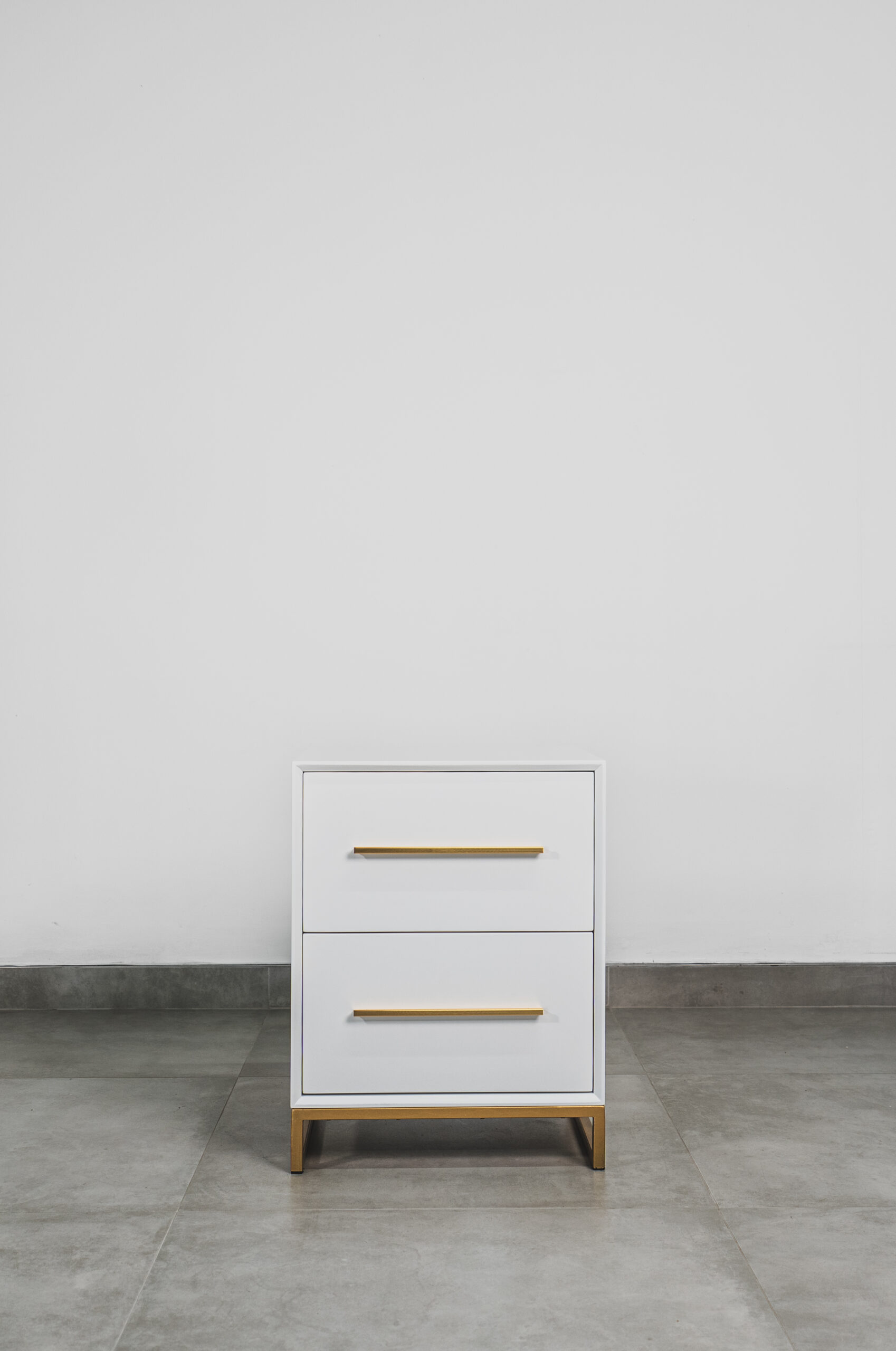 Front view of a modern white bedside table with two drawers, gold handles, and a gold steel base, set on a grey tiled floor against a white wall.