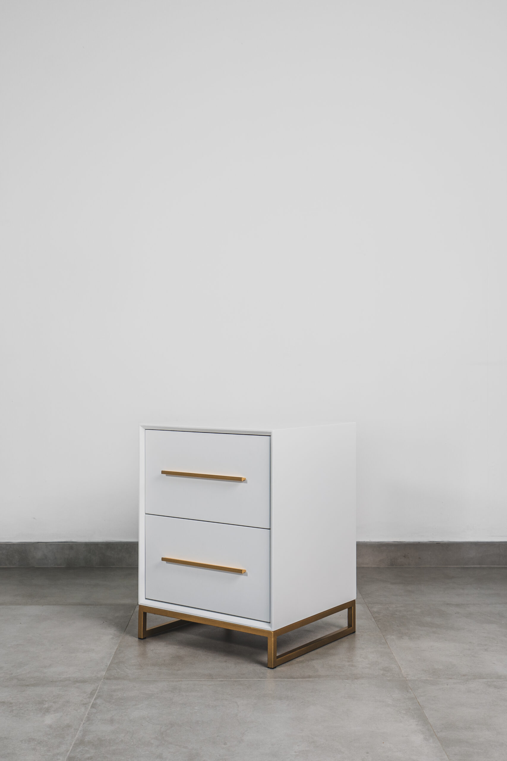 Modern white bedside table with two drawers, gold handles, and a sleek gold steel base on a grey tiled floor.