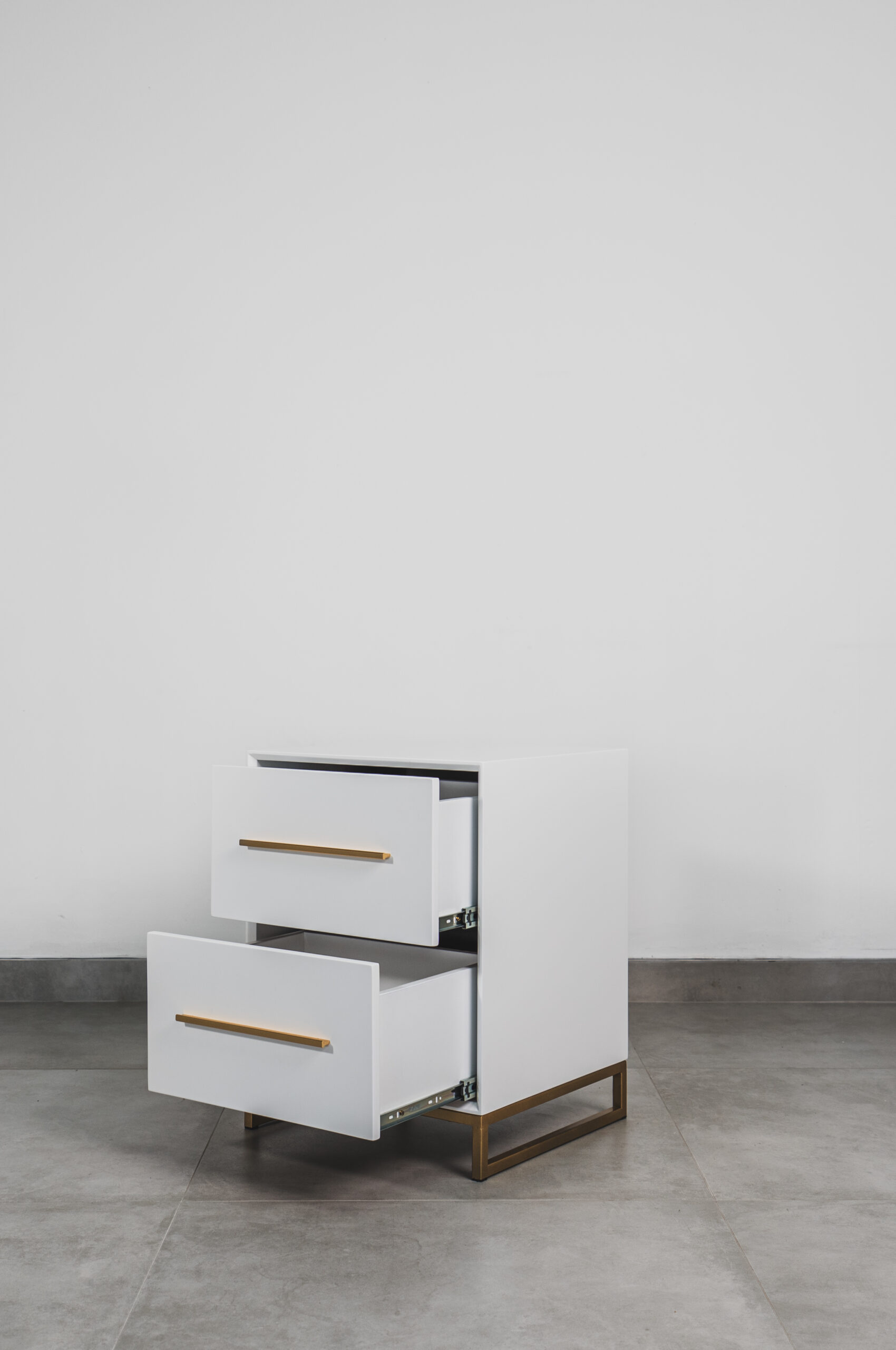 White bedside table with two drawers open, featuring gold handles and a gold steel base, displayed on a grey tiled floor.