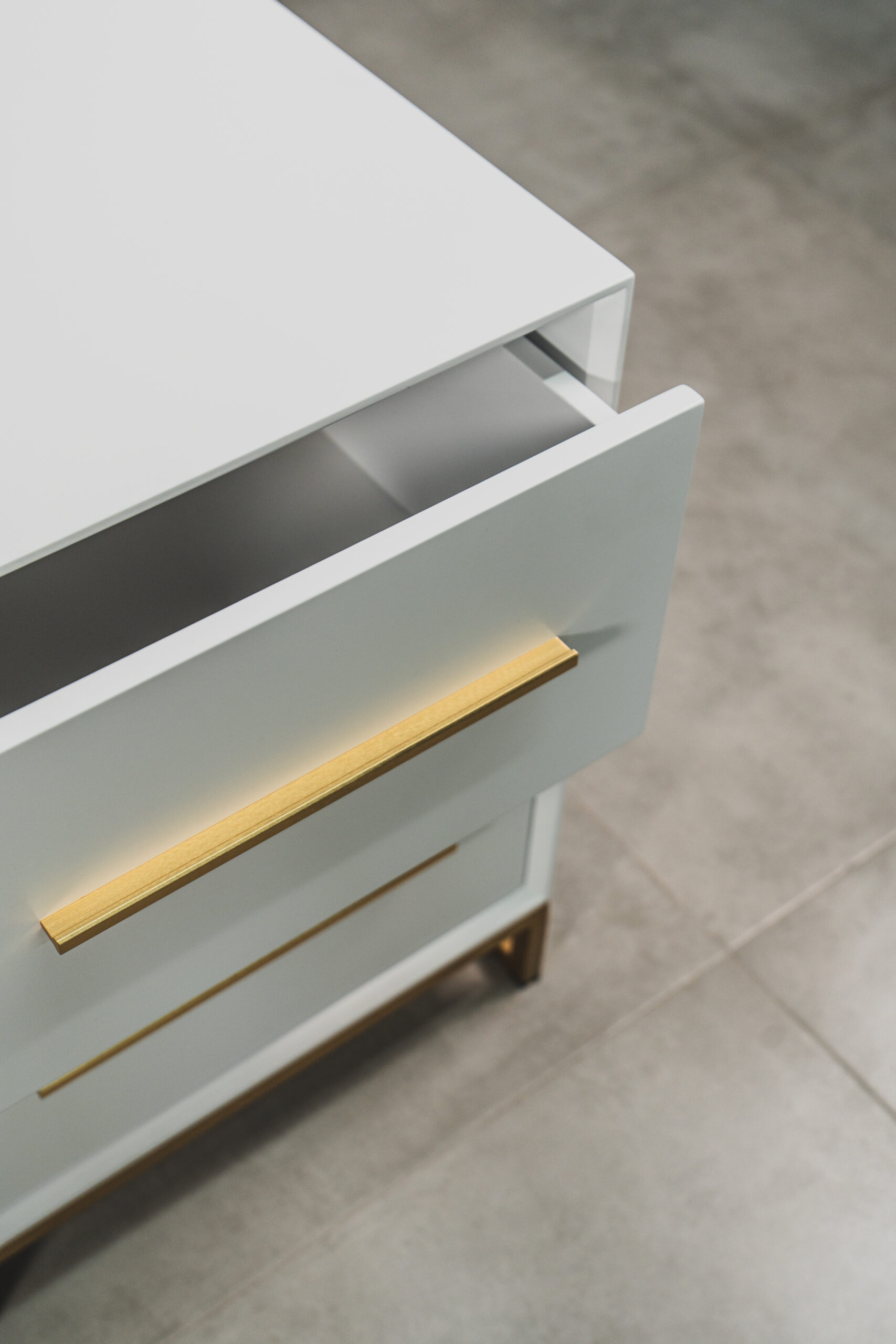Close-up of a modern white bedside table with an open drawer, featuring elegant gold handles and a gold steel base.
