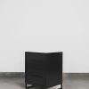 Modern black pedestal with two drawers and a sleek steel base, shown on a grey tiled floor against a white wall.