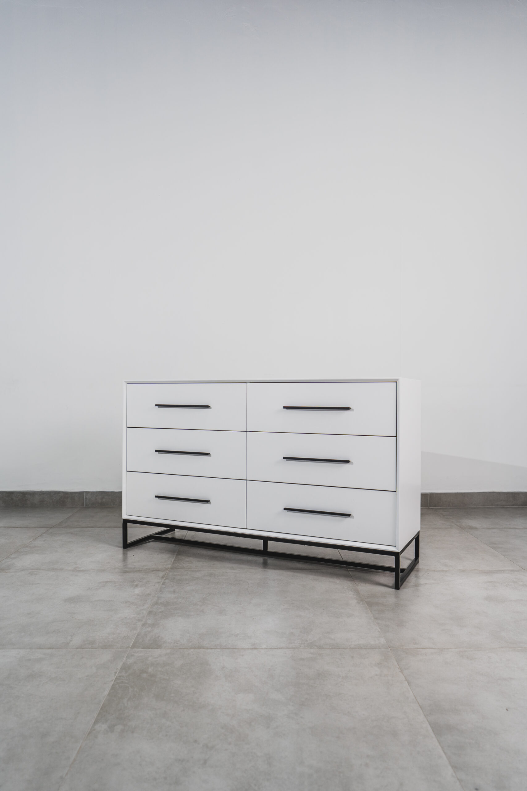 White six-drawer chest of drawers with black handles and steel base on tiled floor, ideal for modern bedroom storage.
