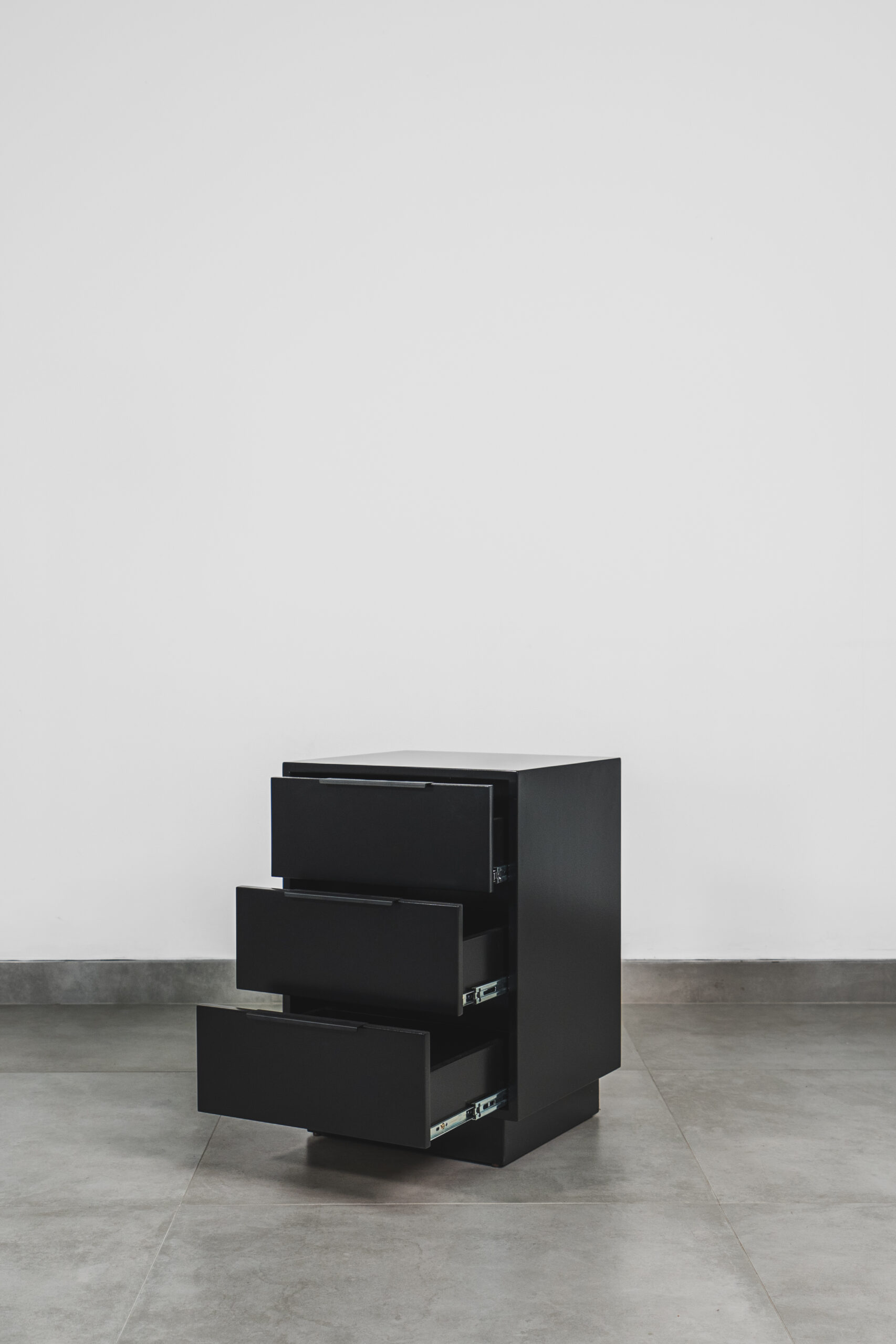 Modern black bedside pedestal with three open drawers, featuring a sleek matte finish and minimalist design.