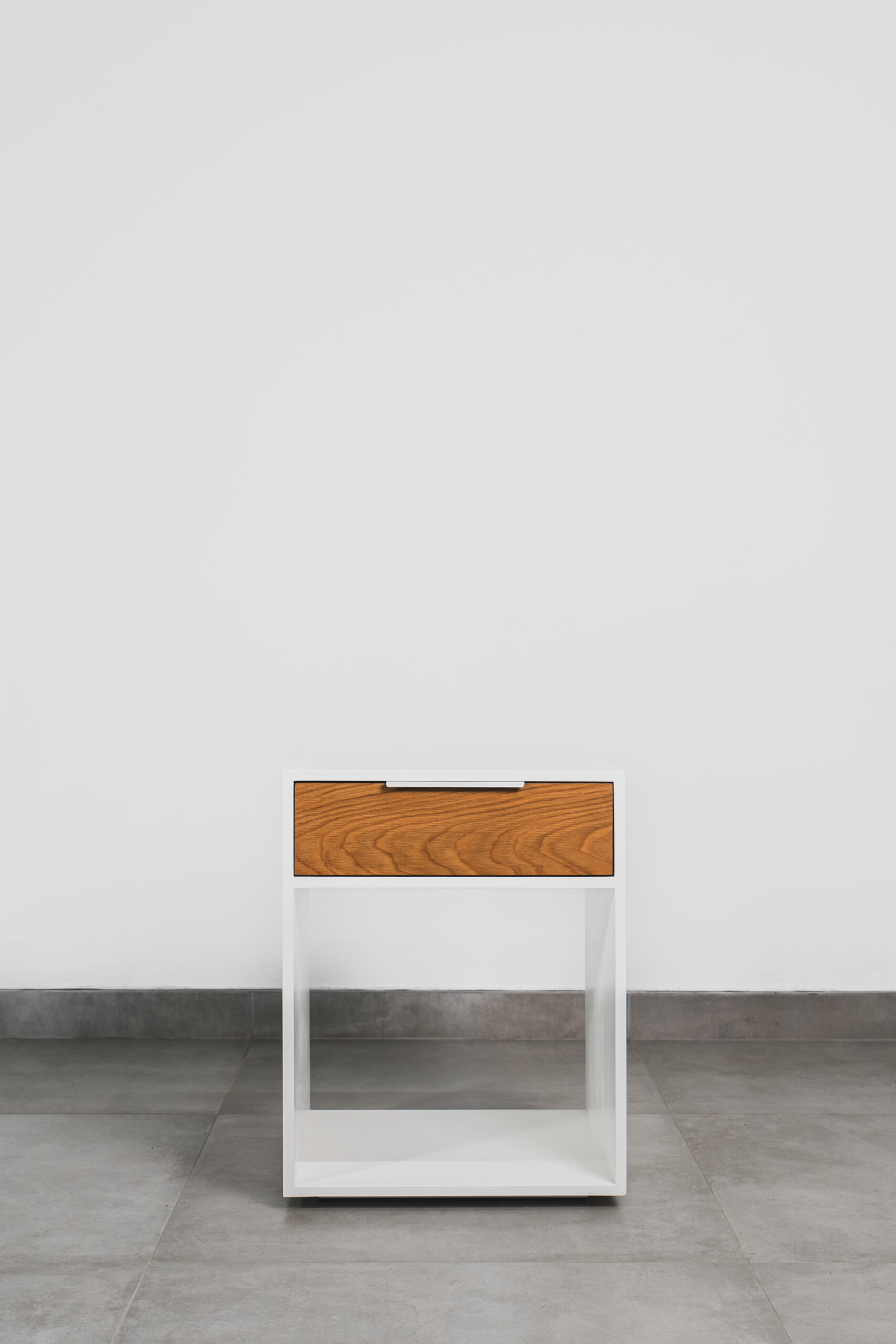 White pedestal with a single wooden drawer and open shelf, featuring a clean, modern design on a tiled floor.
