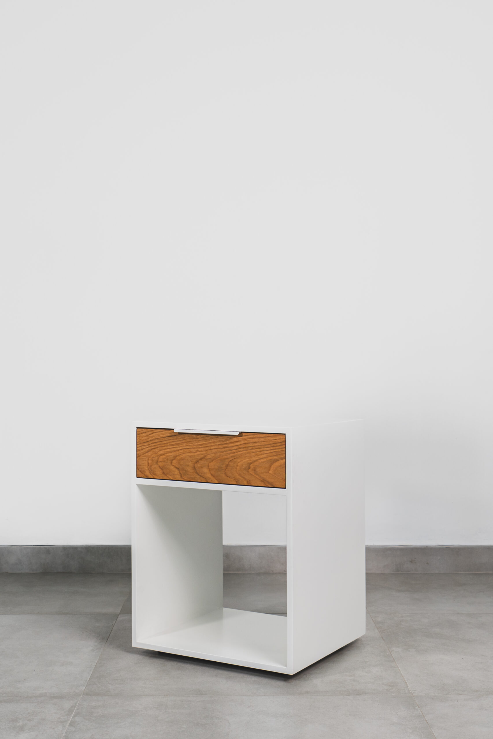 White pedestal with a single wooden drawer and open shelf, featuring a modern minimalist design on a tiled floor.