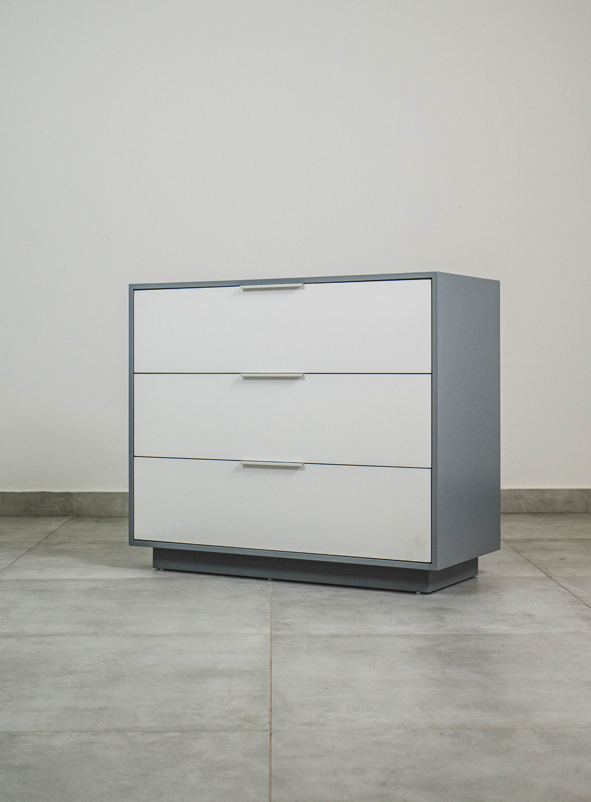 Modern grey and white compactum chest of drawers with three white-front drawers and silver handles, shown angled
